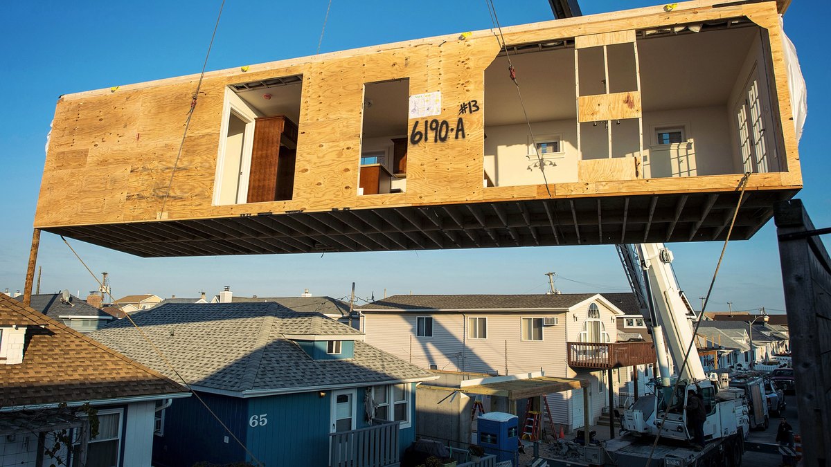A crane lifts a plywood-sheathed modular house section over a street of finished homes